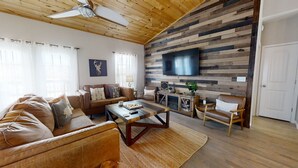 A cozy living room with a wooden ceiling, a large flat-screen TV mounted on a wooden wall, a brown leather sofa, a coffee table, and a fireplace with a TV.