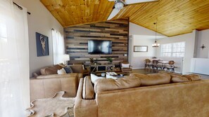 Un salon spacieux avec un grand canapé d’angle, un téléviseur à écran plat fixé à un mur en bois, et un coin repas comprenant une table et des chaises.