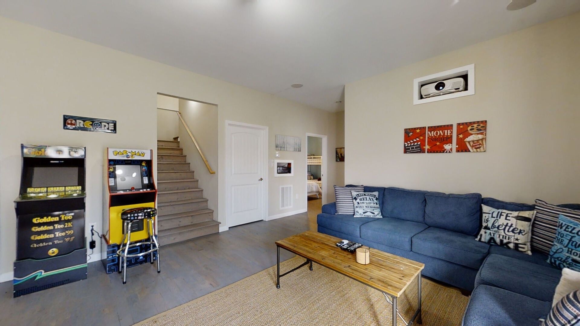 A living room with a blue sofa, a coffee table, arcade games, a staircase, and a door leading to another room.