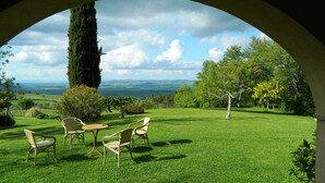 A grassy outdoor area with a table and chairs, a large tree, and a view of distant hills.