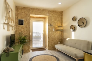 A living room with a beige sofa, a green desk with a telephone, a potted plant, and a wooden door leading outside.