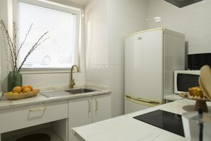 A kitchen with white cabinets, a sink, a window with blinds, a refrigerator, a microwave, and a basket of oranges. - Apartment by the river in Seixal bay. 3pax. Bicycles are available. (Seixal)