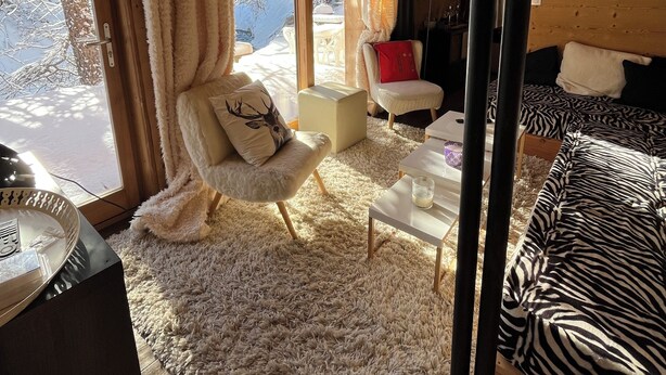 A cozy cabin interior with a wooden ceiling, a plush fur rug, a zebra-patterned bed, a white armchair with a deer pillow, and a dining table with a glass of milk.