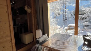 A wooden cabin interior with a dining table covered in plastic, a cushioned bench, and a view of a snowy landscape outside.