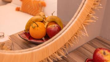 Un espejo con diseño de flecos, que refleja una cama con almohadas blancas y un cabecero de madera. Frente al espejo, hay una mesa de madera con un plato de fruta que incluye plátanos, naranjas y manzanas.