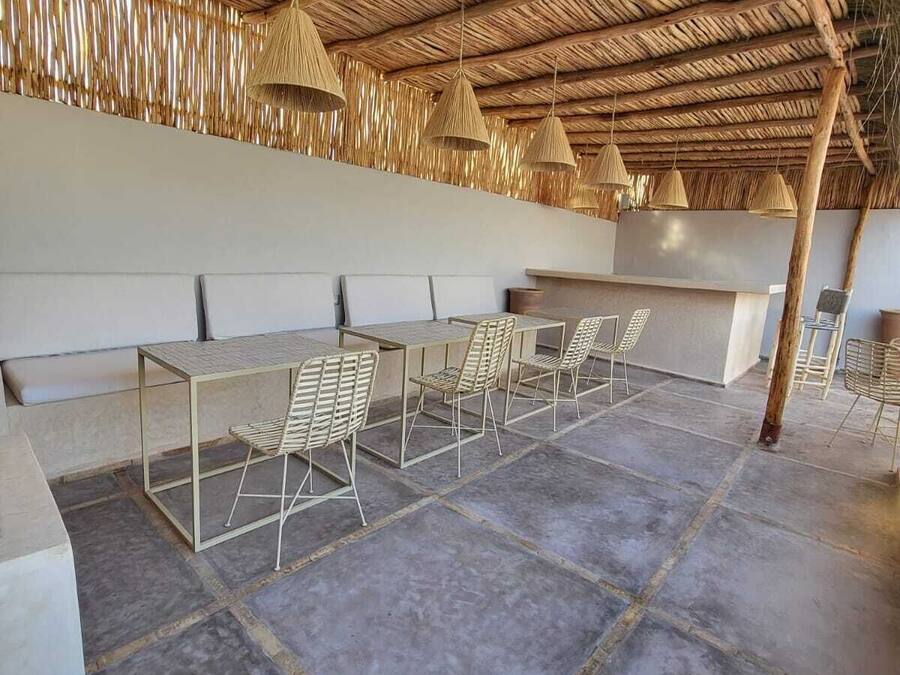 Un área de estar al aire libre con techo de paja, mesas de metal y sillas de mimbre.
