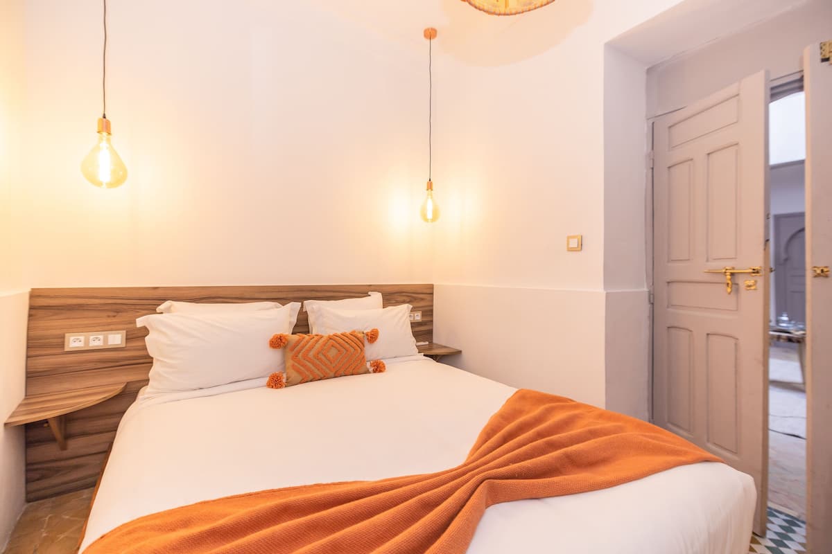 A neatly made bed with white linens and an orange throw blanket, a wooden headboard, and two hanging light bulbs.