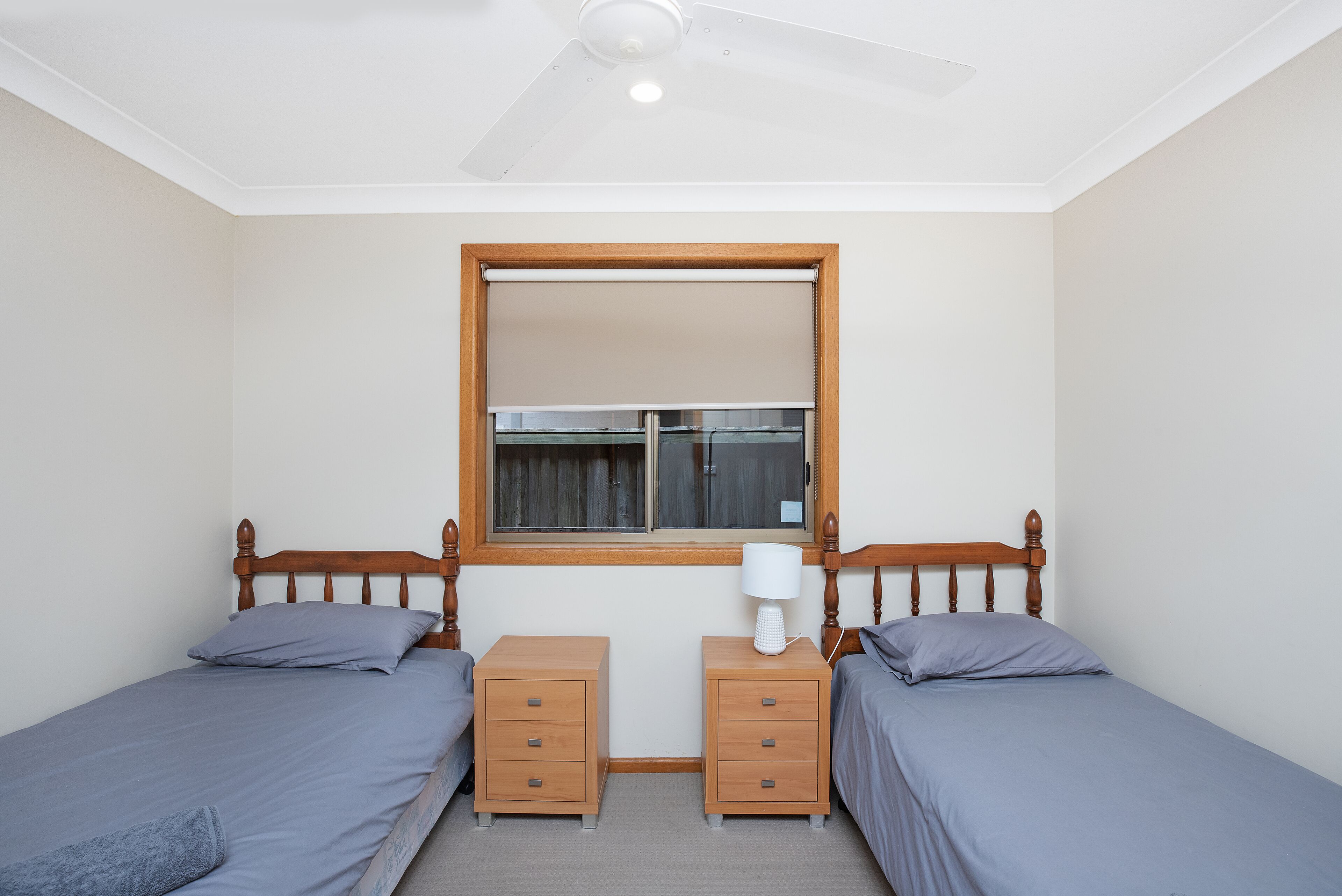 Une chambre avec deux lits simples, des têtes de lit en bois, une fenêtre avec un cadre en bois, un ventilateur de plafond et une table de chevet avec une lampe.