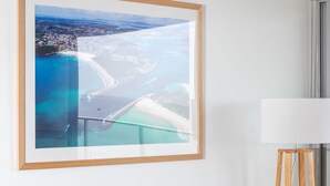 A tan leather sofa with patterned cushions, a framed coastal landscape photograph on the wall, and a floor lamp. - Oceanic 7 - Opposite from main beach (Forster)