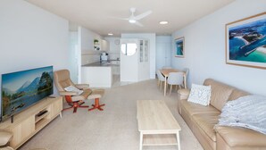 A modern living room with a TV, seating, a dining area, and a kitchen in the background. - Oceanic 7 - Opposite from main beach (Forster)