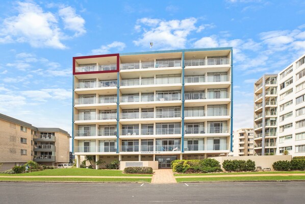 A multi-story hotel with a central entrance, balconies on each floor, and a parking area in front.