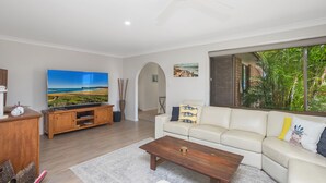 A living room with a flat-screen TV, a wooden entertainment unit, a white sofa with cushions, a coffee table, and a large window with a view of greenery. - Spacious. Tropical. Waterfront. (Tuncurry)