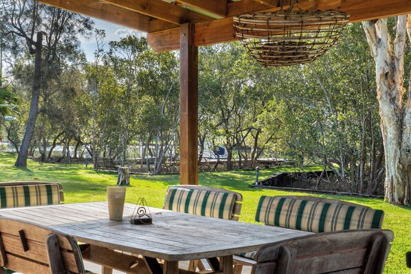 Una terraza exterior de madera con una mesa y sillas, rodeada de árboles y un área con césped.