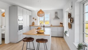 Une cuisine moderne avec des meubles blancs, un îlot central en bois doté de tabourets, un luminaire suspendu et une vue sur l’extérieur grâce à une grande fenêtre.