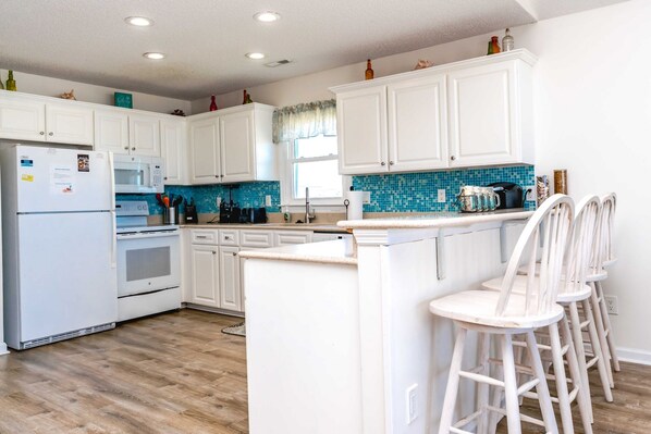 Una cocina con armarios blancos, un refrigerador, un horno, un lavamanos y un salpicadero de azulejos azules. Hay tres taburetes blancos y una ventana con cortina.