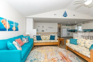 A living room with two sofas, a coffee table, and a rug. There is a kitchen in the background with white cabinets and a dining area.