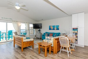 A living room with a wooden sofa, a blue couch, a wooden coffee table, and a dining table with chairs. There is a television on the wall, a ceiling fan, and a view of the ocean through large windows. - Followin The Son - Amazing 4 BR Oceanfront Duplex; Great Views! (North Topsail Beach)