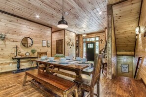 Un intérieur rustique en bois, avec une table à manger pour quatre personnes, des bancs, un lustre et un escalier menant à un autre niveau.