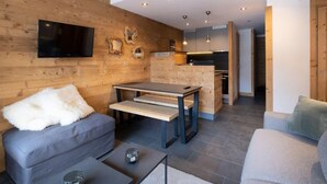 A modern living room with a wooden wall, a grey sofa, a small table, and a kitchen area in the background.