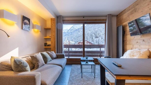 A modern living room with a sofa, a coffee table, and a TV. There is a large window with a view of snow-covered mountains.