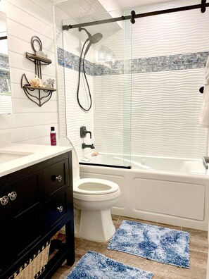 A bathroom with a white toilet, a bathtub, a glass shower enclosure, a vanity with a mirror, and decorative wall shelves.