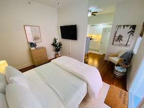 A bedroom with a large bed, a TV mounted on the wall, a wooden dresser, a plant, and a view into the kitchen.