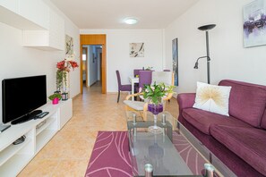 Una sala de estar moderna con un televisor de pantalla plana, una mesa de centro de vidrio, un sofá púrpura y una zona de comedor al fondo.