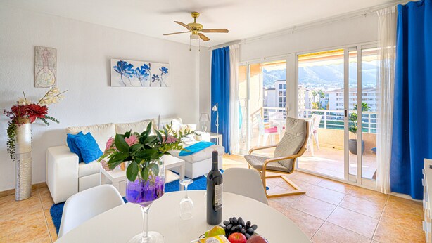 Un salon moderne avec un balcon, une table à manger ornée de fruits et d’une bouteille de vin, et un canapé agrémenté de coussins bleus.