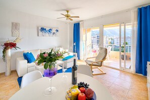 Ein modernes Wohnzimmer mit Balkon, einem Esstisch mit Obst und Wein sowie einem Sofa mit blauen Kissen.