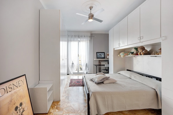 A bedroom with a bed, a ceiling fan, a desk, and a shelf with stuffed animals.