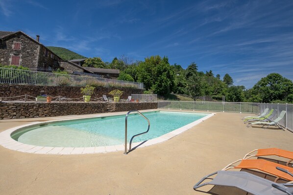 Une piscine avec un escalier en métal, entourée d’une clôture blanche, et un bâtiment en pierre en arrière-plan.
