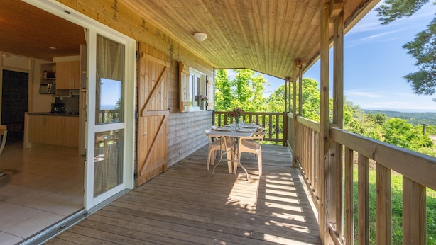 Een houten terras met een tafel en stoelen, een schuifdeur naar een keuken en uitzicht op groen.