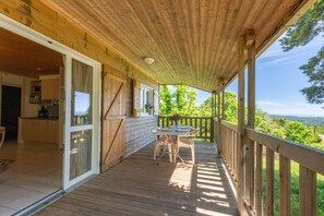 Une terrasse en bois avec une table et des chaises, une porte-fenêtre coulissante donnant sur une cuisine et une vue sur la verdure.