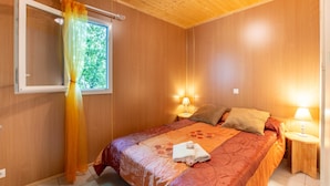 Een kamer met houten wanden, een bed met een bloemenprint, twee bedlampjes, een raam met gordijn en een klein nachtkastje.