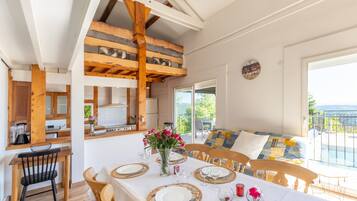 Un coin repas avec une table dressée pour quatre personnes, des poutres en bois, une cuisine en arrière-plan et un balcon avec vue.