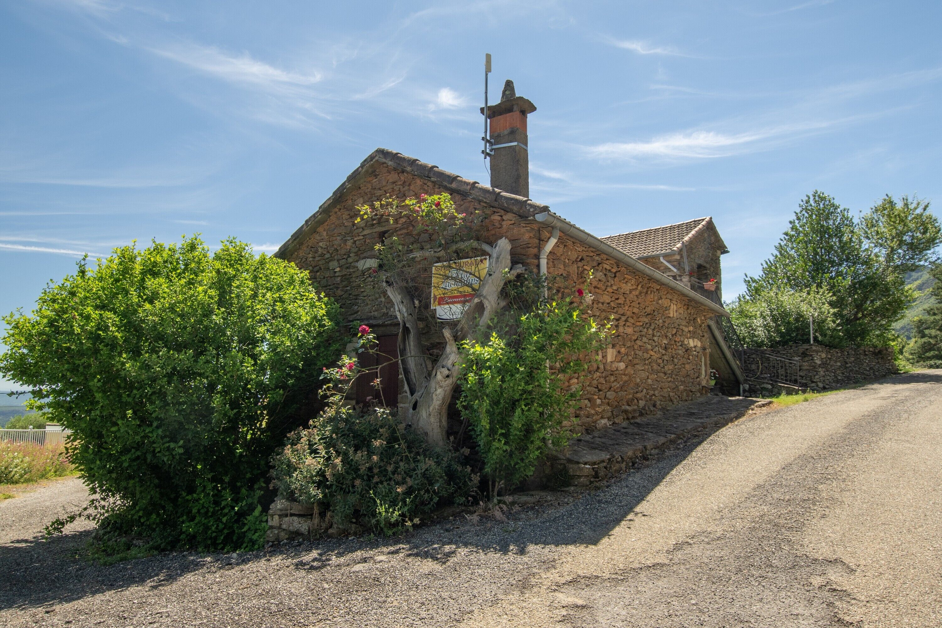 A stone building with a chimney, surrounded by greenery and a gravel path.