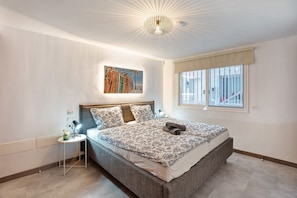 A bedroom with a large bed, a nightstand, a wall-mounted lamp, a ceiling light, a window with a view, and a painting on the wall.