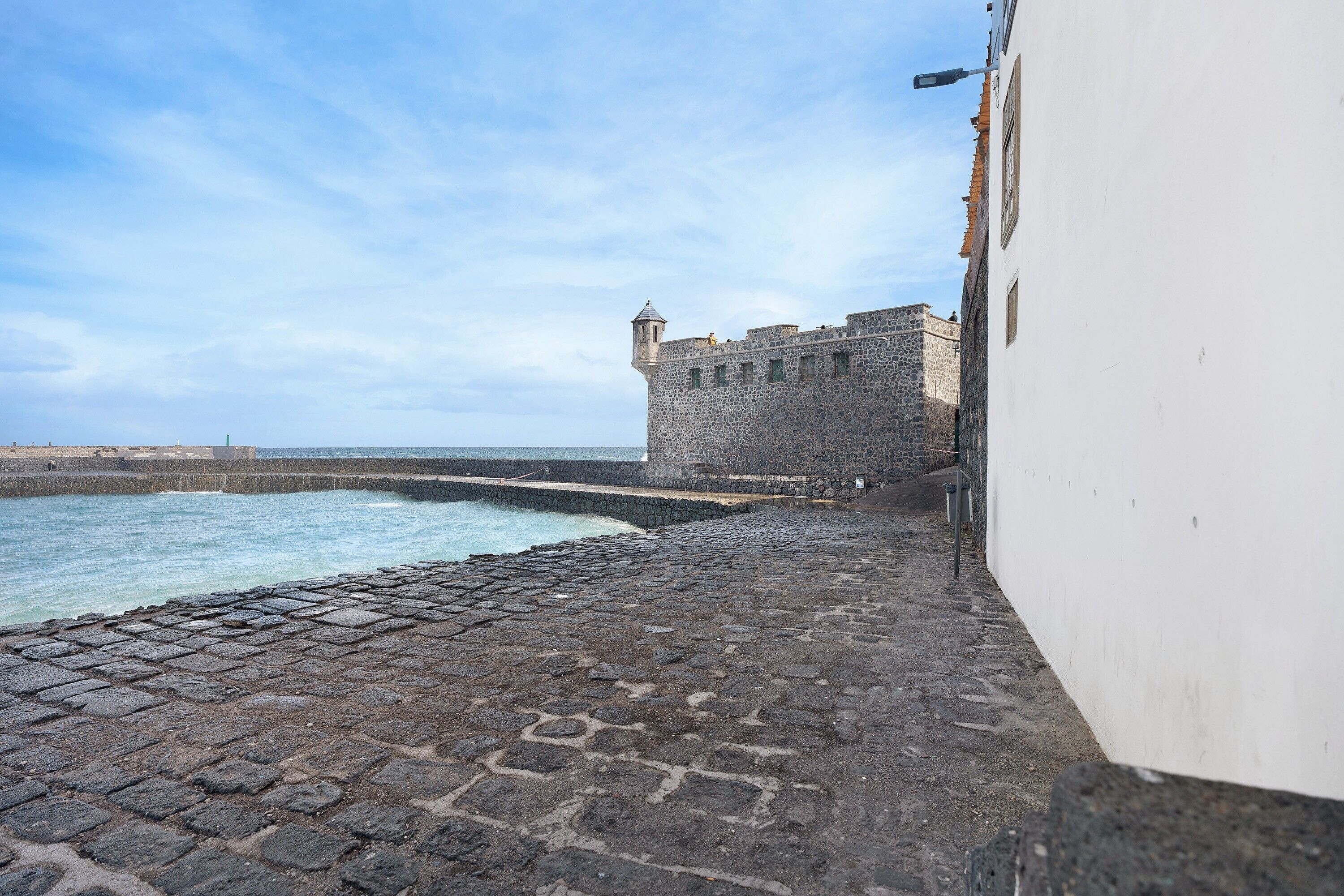 Un sendero adoquinado conduce a un edificio histórico de piedra con una torre, con vistas al mar tranquilo.