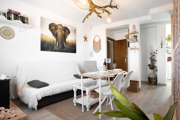 A cozy living area with a white sofa, a dining table with chairs, a large elephant painting, and a unique ceiling light fixture.