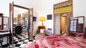 Ein Hotelzimmer mit Schachbrettmuster auf dem Boden, einem Bett mit einer rot-beigen Steudecke, einem Holzstuhl, einem Schreibtisch mit Stuhl und einem Fernseher.