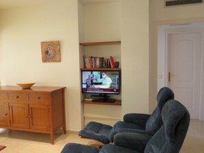 A living room with a television, a wooden cabinet, a blue armchair, and a bookshelf.