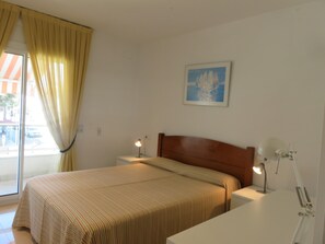 A hotel room with a bed, bedside tables, a lamp, a painting on the wall, and a window with curtains. - Apartment "Brises del Mar 1-1" close to the Sea with Shared Pool & Wi-Fi (Girona)