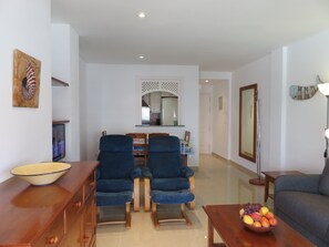 A living room with a sofa, two armchairs, a wooden coffee table with fruit, a TV, and a decorative wall piece.