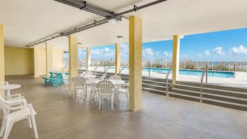 Un espace extérieur abrité, doté d’un sol en béton, de chaises et de tables en plastique blanc, et offrant une vue sur une piscine.