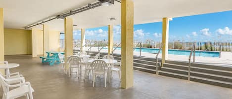 A covered outdoor area with concrete flooring, white plastic chairs, and tables, overlooking a swimming pool.