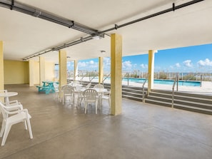 A covered outdoor area with concrete flooring, white plastic chairs, and tables, overlooking a swimming pool. - Shoreline Towers (Gulf Shores)
