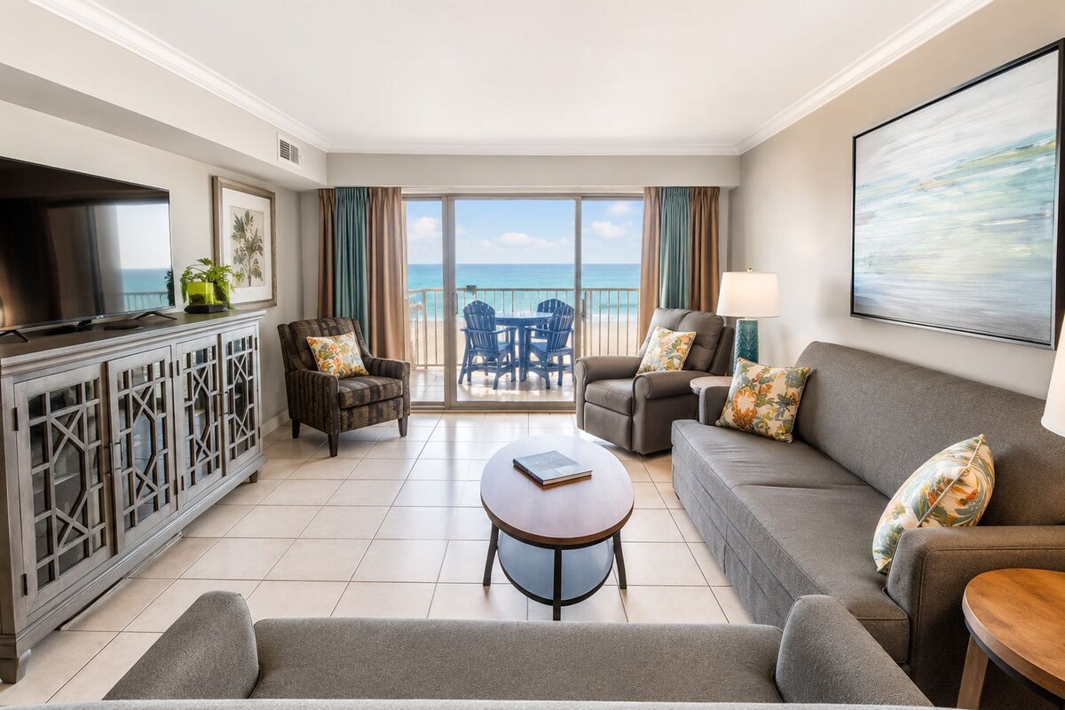 A modern living room with a sofa, armchairs, a coffee table, and a TV on a cabinet. There is a balcony with a view of the ocean.