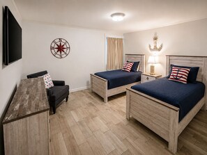 Quarto de hotel com duas camas de madeira, uma escrivaninha, uma cadeira, televisão e arte decorativa de uma bússola na parede.