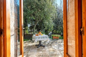 Une porte en bois ouverte donne sur un patio extérieur où une table est dressée avec des fruits et un panier. L’ensemble est entouré d’un sol en pierre et de verdure.