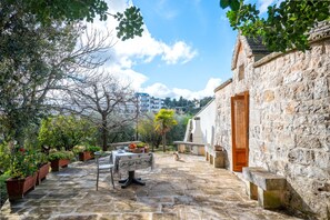 Une cour dallée avec une table et des bancs, entourée de plantes en pot et d’arbres.